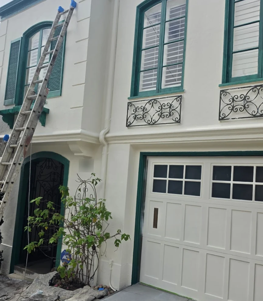 The same house front after painting: dark green trim, even white walls and a white garage door, with a ladder