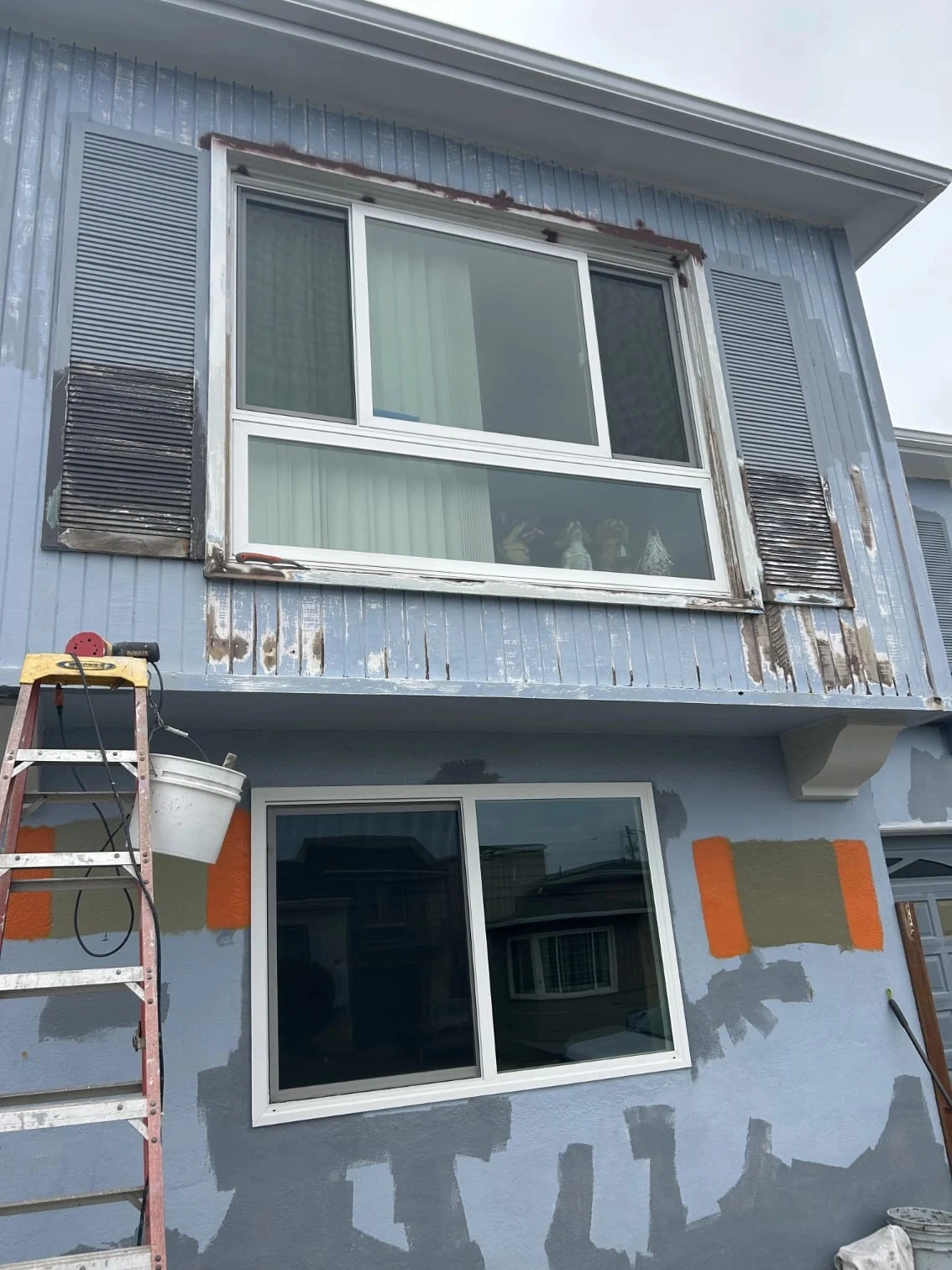 Blue house front during paint preparation, with sanded trim, primer patches and a ladder