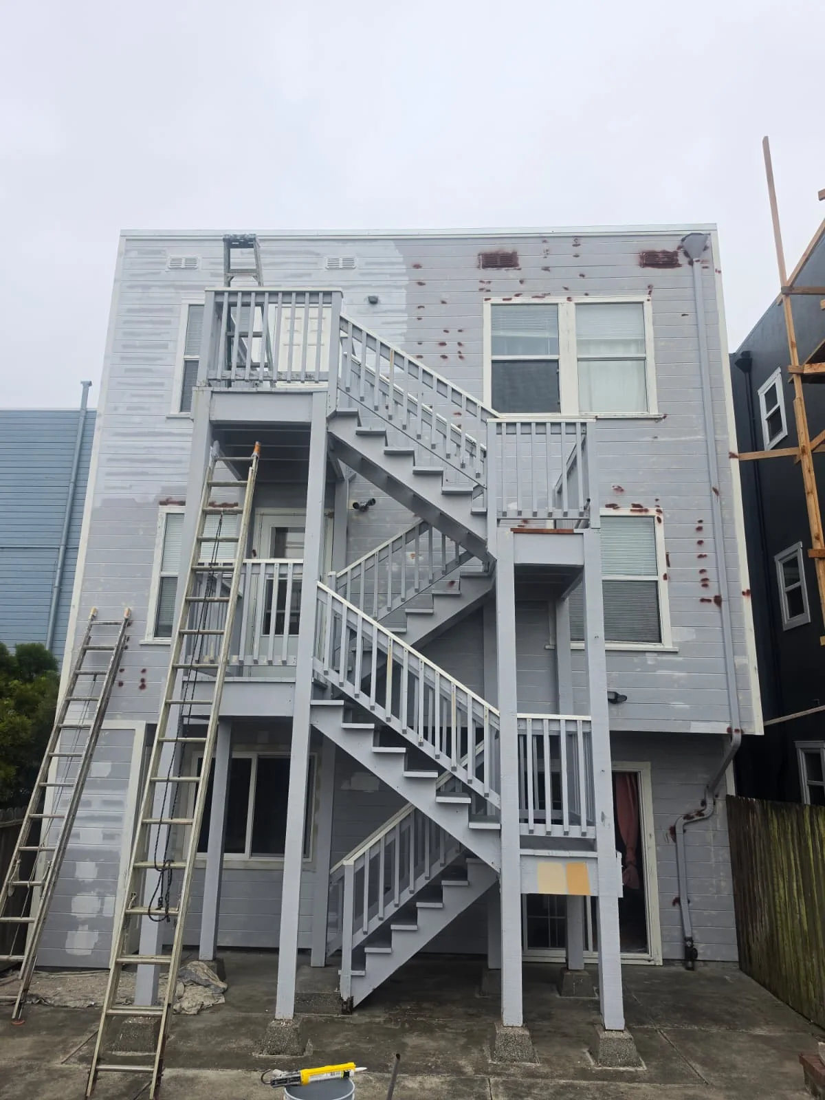 Back stairs and gray-blue siding before painting, with primer patches and ladders